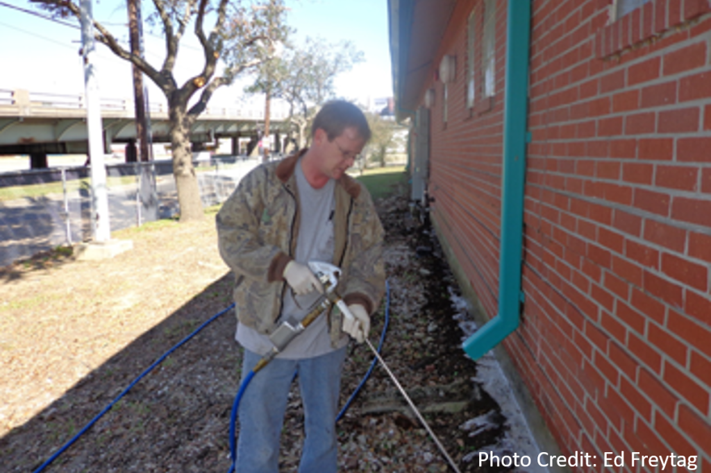 A man is standing outside of a brick building, spraying the outside of the building. He is wearing latex-style gloves.